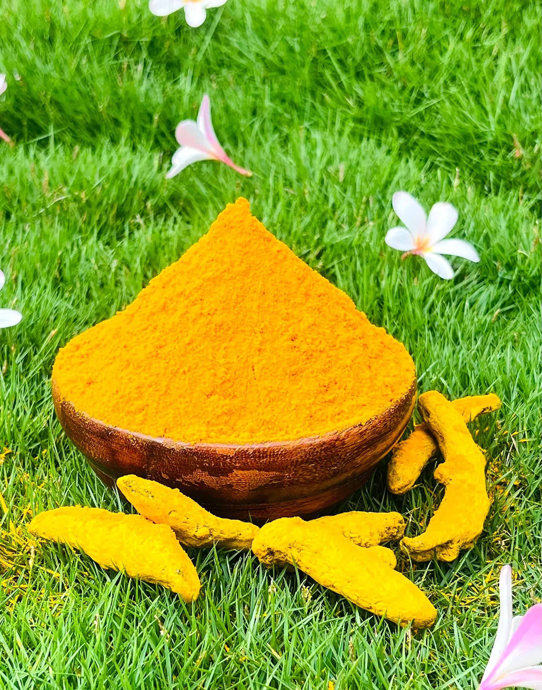 A cone of turmeric powder in a wooden bowl set on grass, frangipani flowers scattered around it