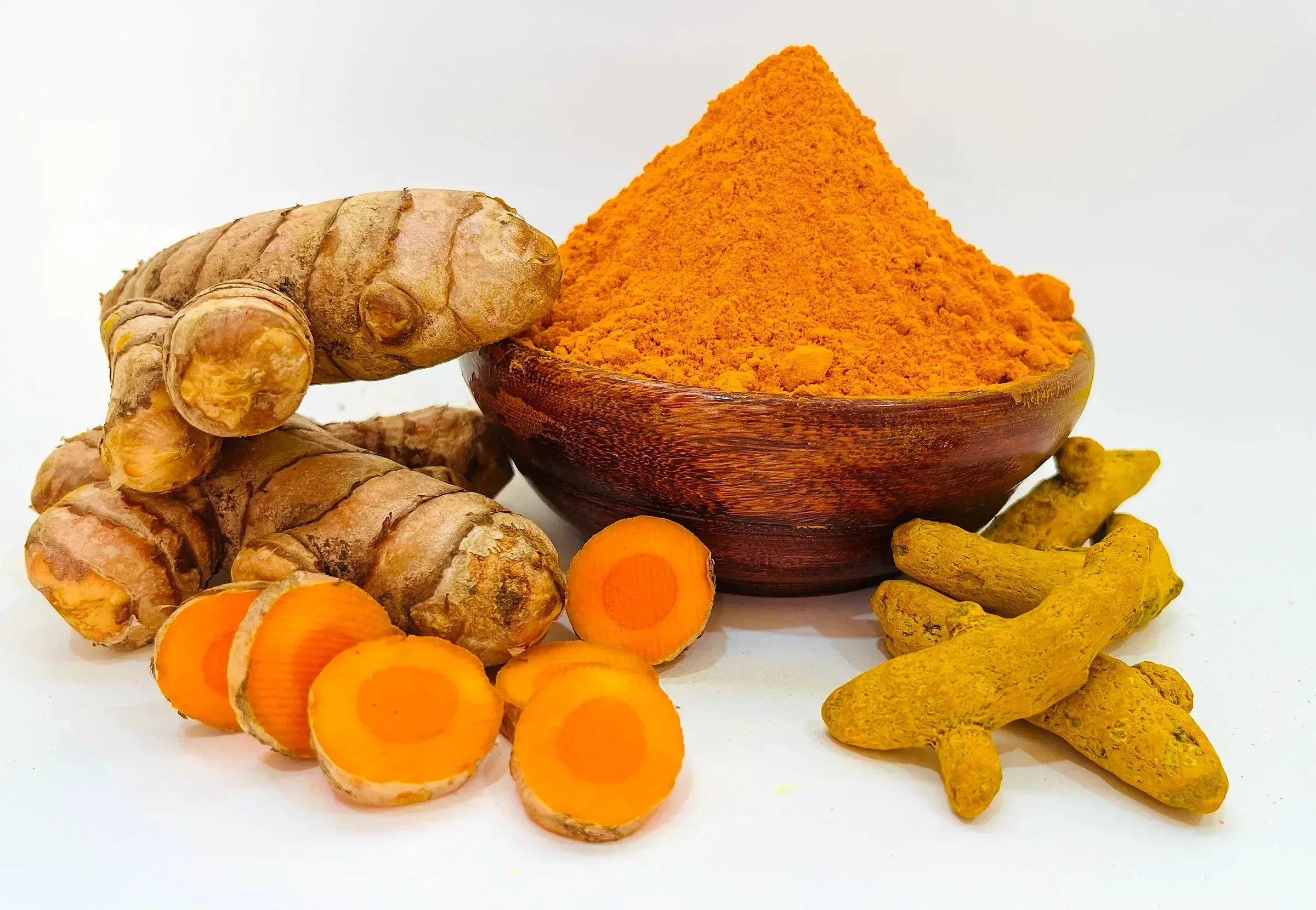 Turmeric in every form on a white ground — fresh rhizomes, cut slices, dried fingers and a wooden bowl heaped with ground powder