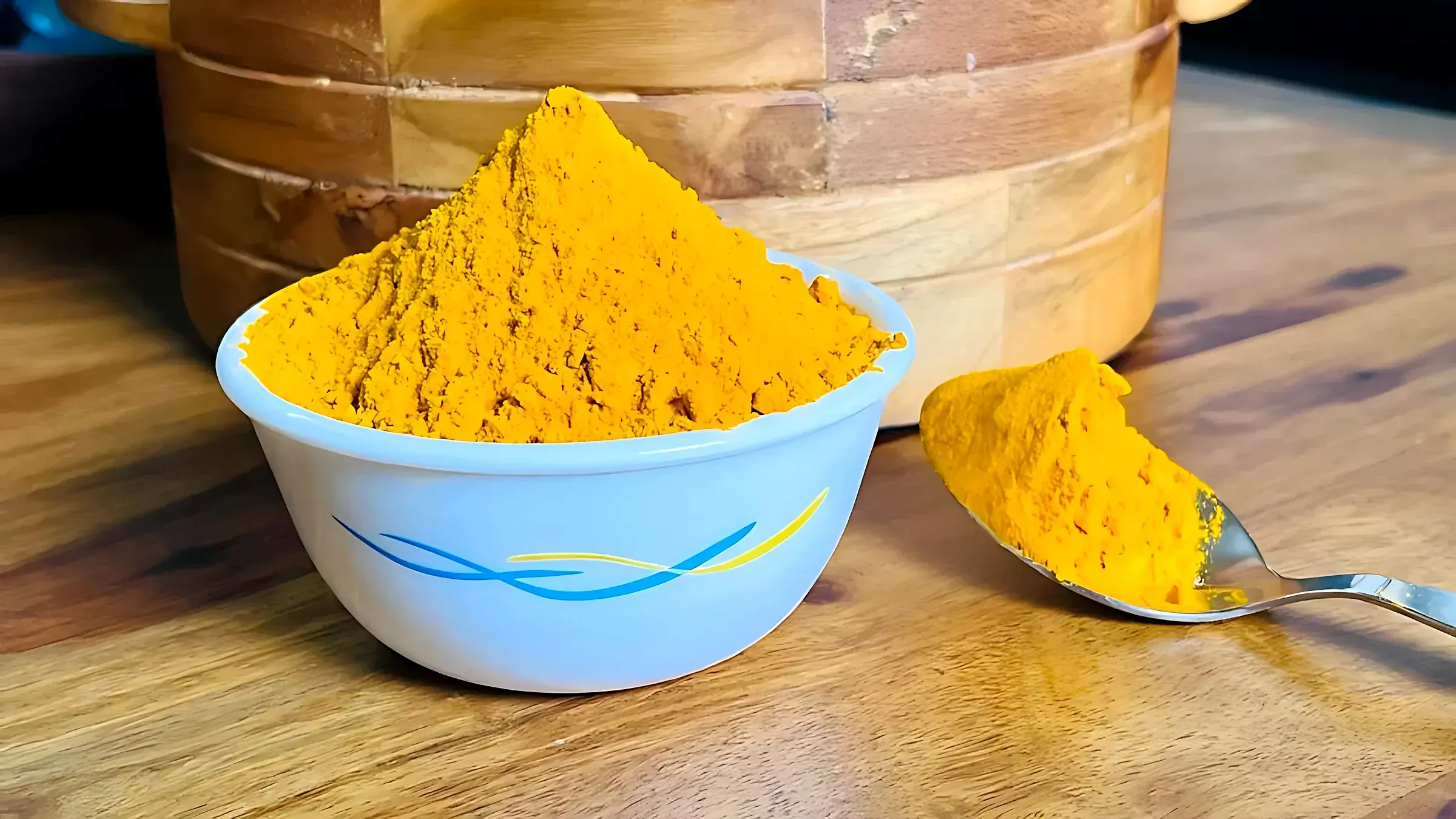 Turmeric powder heaped in a white bowl beside a full spoon on a wooden table
